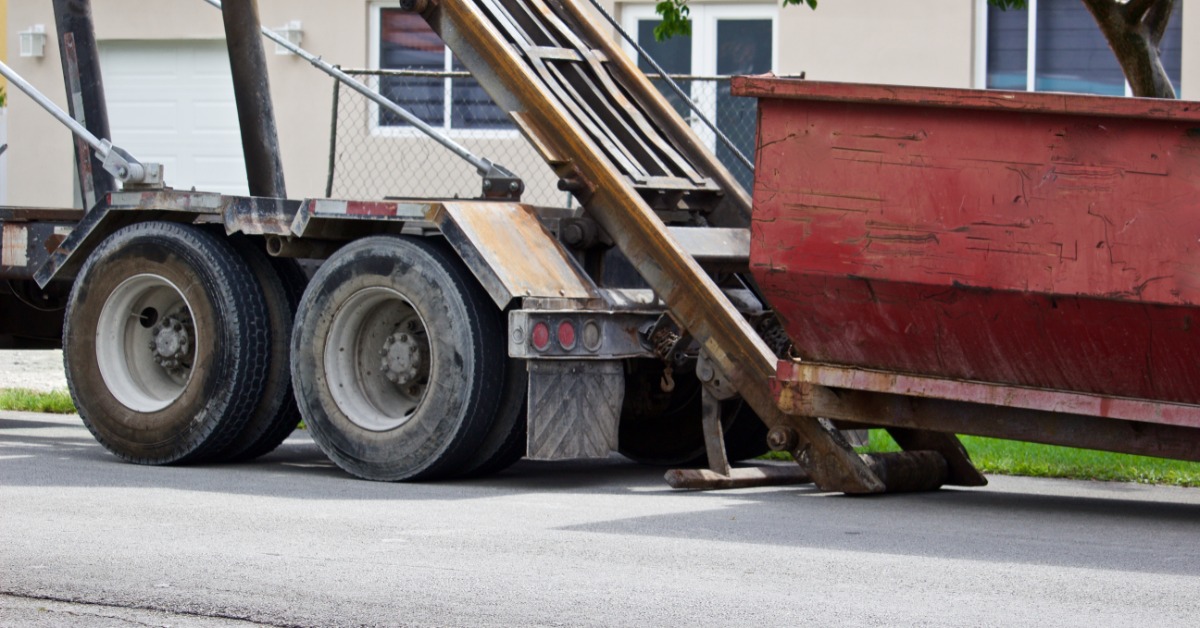 A truck with long railings in the back dropping off a red dumpster on the side of a street in front of a small building.